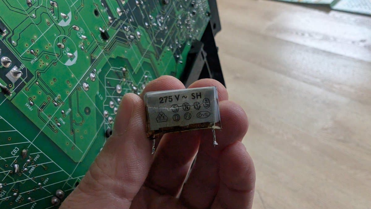 Damaged X2 safety capacitor labeled "275 V ~ SH" held in fingers, showing catastrophic failure with exposed internal metal components.