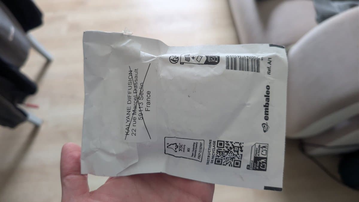White mailing envelope held in hand, addressed to Emmaüsleo in France, with shipping label and QR code visible.