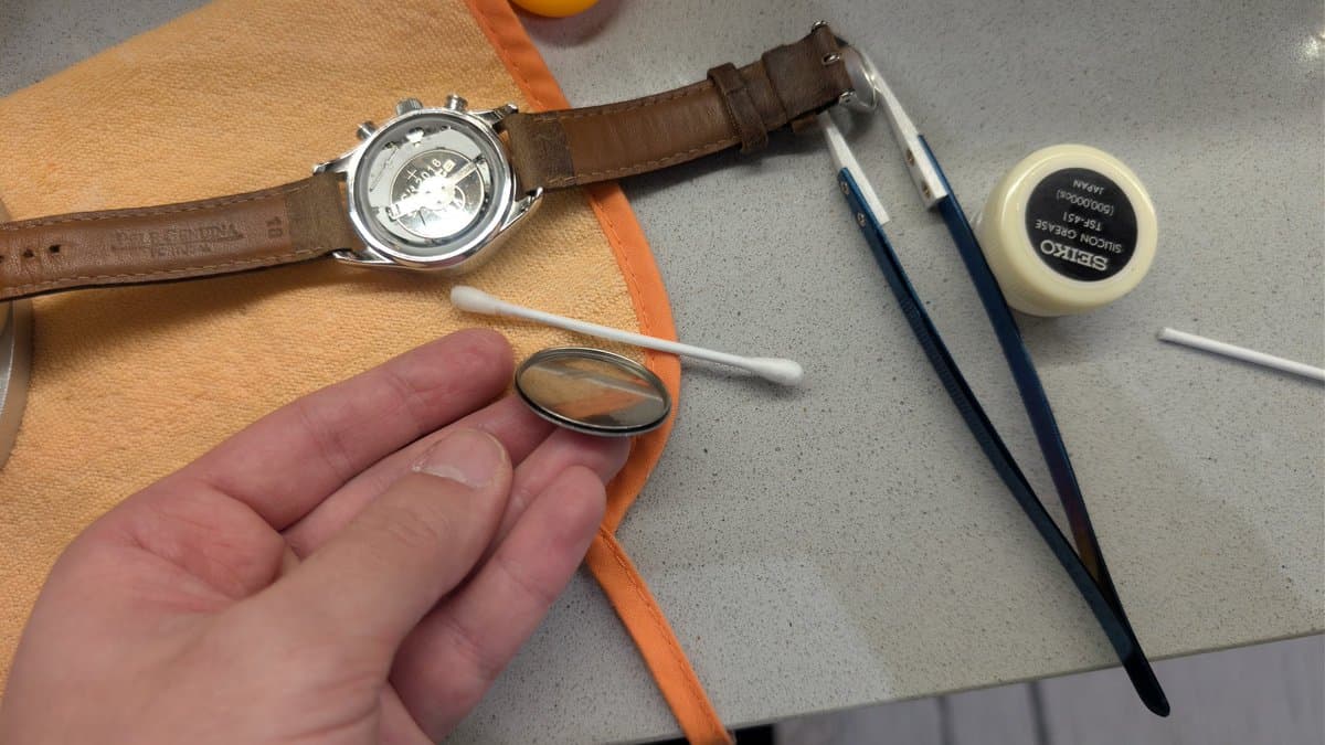 Watchmaking tools and components: brown leather strap, chronograph watch, magnifying glass, brushes, and Breitling gasket lube during battery replacement service.